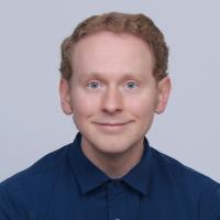 A portrait of a man with red hair and blue eyes wearing a dark blue button-up shirt smiling for a portrait in front of a light blue background.